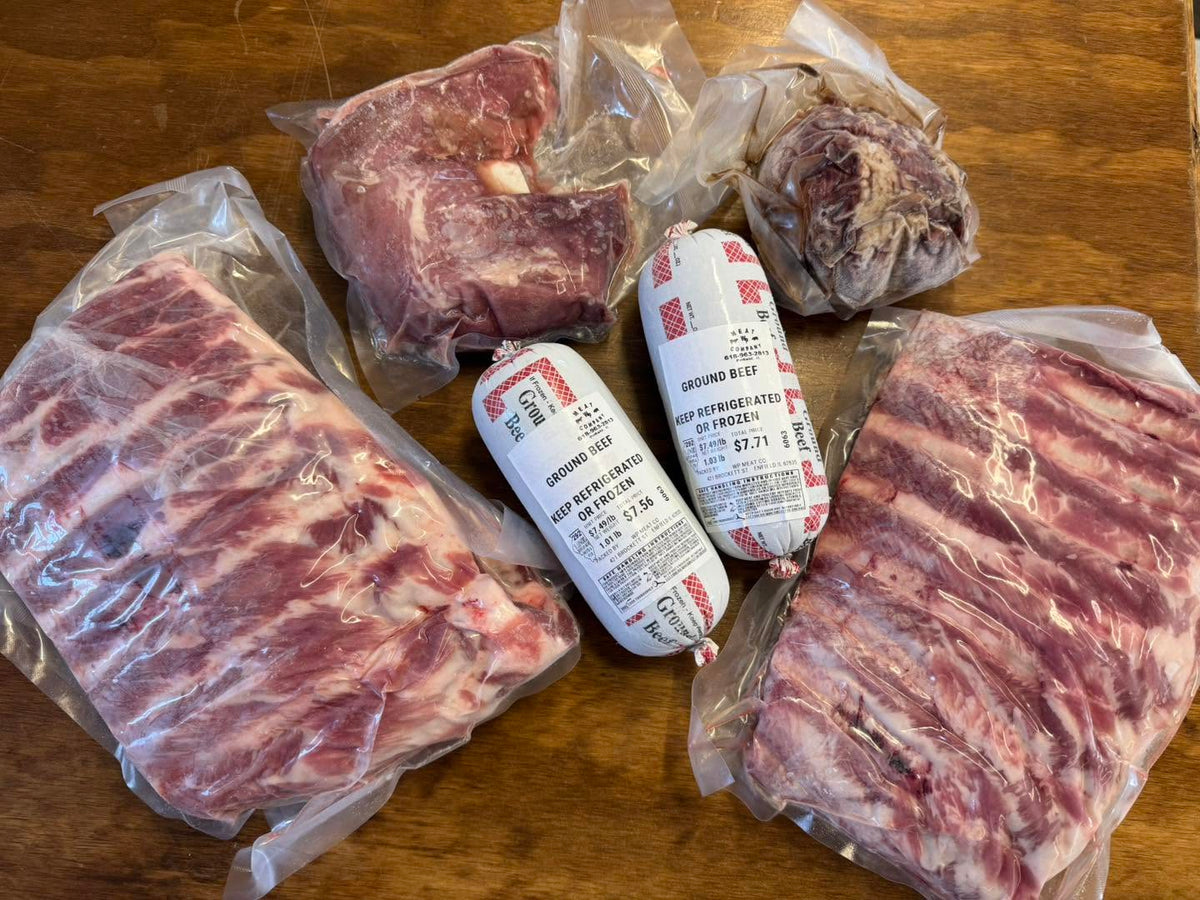 Frozen meat products in clear plastic vacuum packaging arranged on a wooden surface, including a large rib section, another cut of meat, two white-and-red packaged sausages, and a smaller round piece of meat offered at WP Meat Co in Enfield, Illinois.