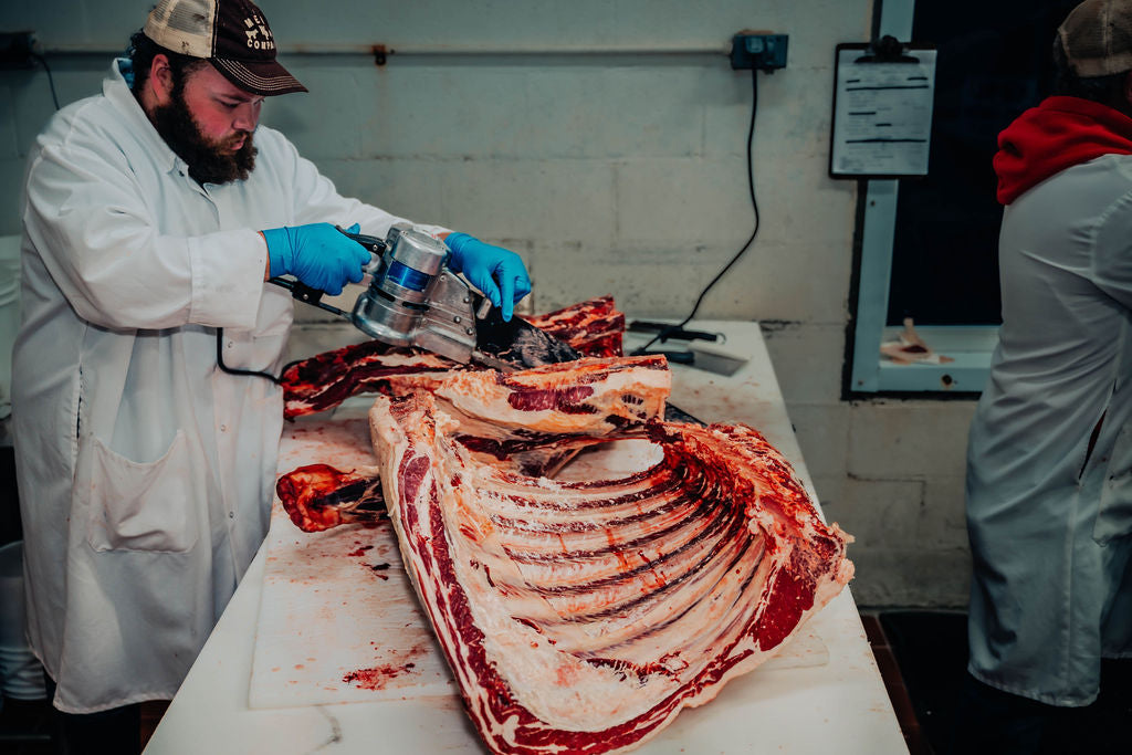 A person in a white butcher coat and blue gloves uses an electric meat knife to process a large rib section at WP Meat Co in Enfield, Illinois.